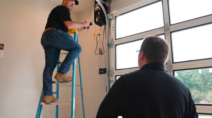 In a busy garage, one person stands on a ladder expertly working on electrical equipment for the commercial overhead doors, while the other carefully observes from the ground.